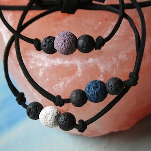 May include: Three black cord bracelets with lava rock beads. The beads are in different colors: white, purple, and blue.