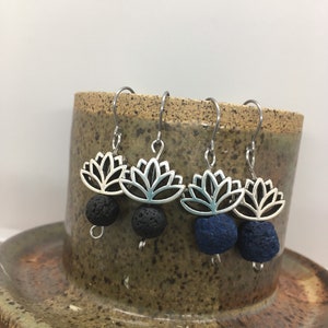 Puede incluir: Pendientes plateados con forma de flor de loto y cuentas de piedra volcánica negra y azul. Los pendientes se exhiben en una taza de cerámica. El diseño de la flor de loto es abierto y detallado, y las cuentas de piedra volcánica tienen una textura.