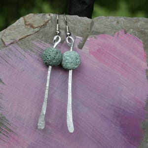 Puede incluir: Pendientes hechos a mano con una cuenta esférica texturizada de color verde claro y una barra larga de plata martillada. Los pendientes cuelgan de ganchos para la oreja de plata. El fondo es una superficie pintada de color púrpura y gris.