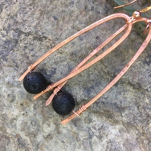 Puede incluir: Pendientes de cobre con un diseño único. Cada pendiente presenta una cuenta de lava negra suspendida de un marco de alambre de cobre. El cobre tiene una textura martillada, lo que añade interés visual. Los pendientes se muestran sobre una superficie de piedra natural.