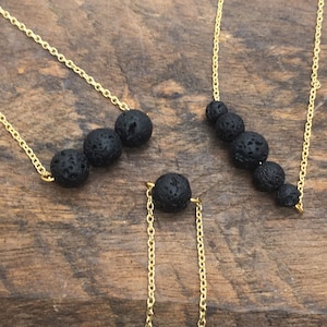 May include: Several gold-colored chain necklaces with black lava rock beads. The necklaces are arranged on a wooden surface. The lava rock beads are round and have a porous texture. The chains are delicate and shiny.