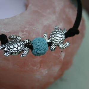 Könnte beinhalten: Ein schwarzes Kordelarmband mit drei silbernen Schildkrötenanhängern und einer blauen Lavakugel.