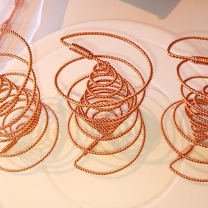 Puede incluir: Tres esculturas de alambre de cobre en forma de espiral. Las esculturas están dispuestas en fila sobre una superficie blanca.