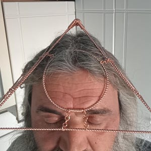 Può includere: Un copricapo a forma di piramide in filo di rame con una fascia circolare intorno alla fronte. Il copricapo è indossato da una persona con i capelli grigi.