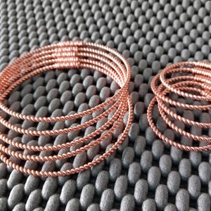 Könnte beinhalten: Ein Set aus sechs roségoldenen, gedrehten Drahtarmbändern. Die Armbänder sind übereinander gestapelt.