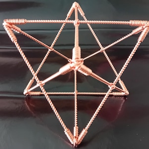 May include: A copper-colored geometric sculpture, featuring a star-like shape with interconnected triangular and rectangular forms. The sculpture is made of twisted wire and sits on a reflective black surface. The overall design is symmetrical and intricate.