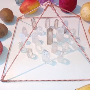 Op de afbeelding: Een piramide van koperdraad met een kristalrooster erin. De piramide staat op een wit oppervlak met fruit eromheen.