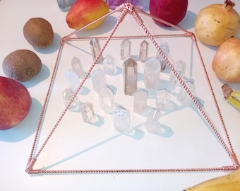Op de afbeelding: Een piramide van koperdraad met een kristalrooster erin. De piramide staat op een wit oppervlak met fruit eromheen.