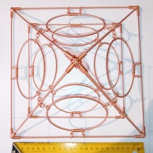 May include: A copper wire pyramid with multiple circular rings, creating a geometric pattern. The pyramid is on a white surface with a ruler in the bottom of the image.