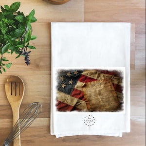 May include: White kitchen towel featuring a distressed American flag and the Declaration of Independence. The towel has a black border around the flag and document. The text "Est. 1776" is printed below the image. Kitchen decor.