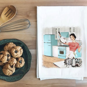 May include: A white kitchen towel with a vintage illustration of a woman in a red shirt and white apron holding a frying pan in a blue and white kitchen. The text "I'm a little teapot" is printed on the towel.