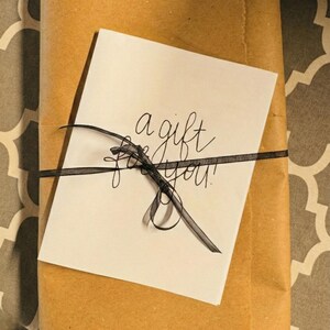 A gift wrapped in brown paper with a black ribbon tied around it, and a white card with black text on top of the paper.