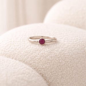 May include: A silver-toned metal ring featuring a single, small, round, deep pink gemstone set in the center. The band is smooth and polished, with a simple, minimalist design that highlights the vibrant color of the circular stone setting.