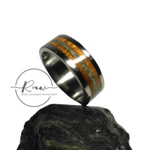 May include: A silver ring with two bands of wood inlay. The wood is a light brown with darker brown streaks. The ring is sitting on a dark gray rock.