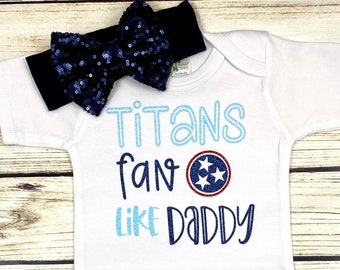 May include: White baby onesie with blue and light blue text that reads "Titans fan like Daddy" with a red and blue Tennessee Titans logo. A blue sequin headband is included.