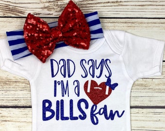 May include: White baby onesie with blue glitter text that says "Dad says I'm a Bills fan" and a red glitter heart with a football inside. The onesie has a red and white striped headband with a red sparkly bow.