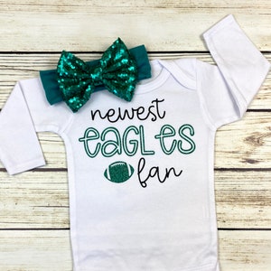 May include: White long-sleeved onesie with the text "newest eagles fan" and a green football graphic. A green sequined bow is attached to the onesie.