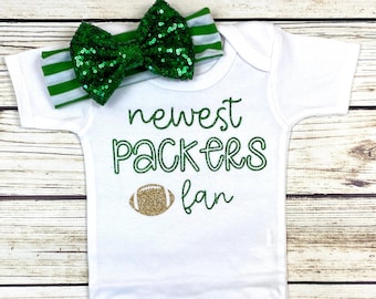 Może przedstawiać: Biały śpioszek dla niemowlęcia z zielono-białą opaską na głowę. Śpioszek ma złoty brokatowy napis "newest Packers fan" z grafiką złotego brokatowego piłki nożnej.
