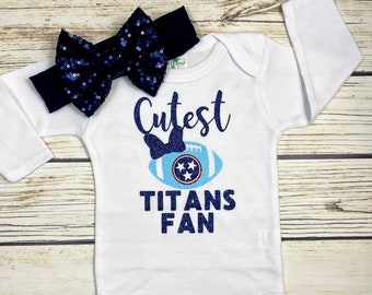 May include: White long-sleeved baby onesie with blue glitter text that reads "Cutest Titans Fan" and a blue glitter football with a blue bow and a Tennessee Titans logo. A blue glitter bow headband is included.