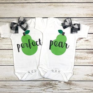 May include: Two white baby onesies with short sleeves. One onesie has a green pear with the text "perfect" above it. The other onesie has a green pear with the text "pear" above it. Both onesies have black and white checkered bows.