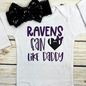 Peut inclure: Combinaison blanche pour bébé avec un texte en paillettes violettes qui dit "Ravens fan like Daddy" avec un ballon de football noir et blanc dans un cœur violet avec un nœud noir. Un bandeau noir à paillettes est sur le dessus de la combinaison.
