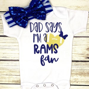 May include: White baby onesie with blue glitter text that says "Dad says I'm a Rams fan". A gold glitter football in a heart shape is above the text. A blue sequined bow with a blue and white striped headband is on top of the onesie.