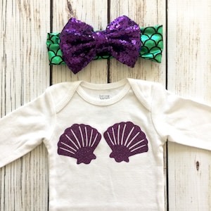 May include: A white baby onesie with purple glitter seashells on the front. A green and purple sequined headband with a bow is also included.