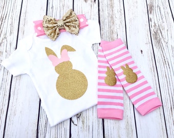 May include: A white onesie with a gold glitter bunny silhouette and pink bow. The onesie is paired with pink and white striped leg warmers with gold glitter bunny accents and a pink and white polka dot headband with a gold glitter bow.
