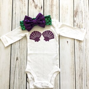 Peut inclure: Combinaison blanche pour b&eacute;b&eacute; avec des coquillages violets sur le devant. Un bandeau &agrave; sequins vert et violet avec un n&oelig;ud est plac&eacute; sur la combinaison.