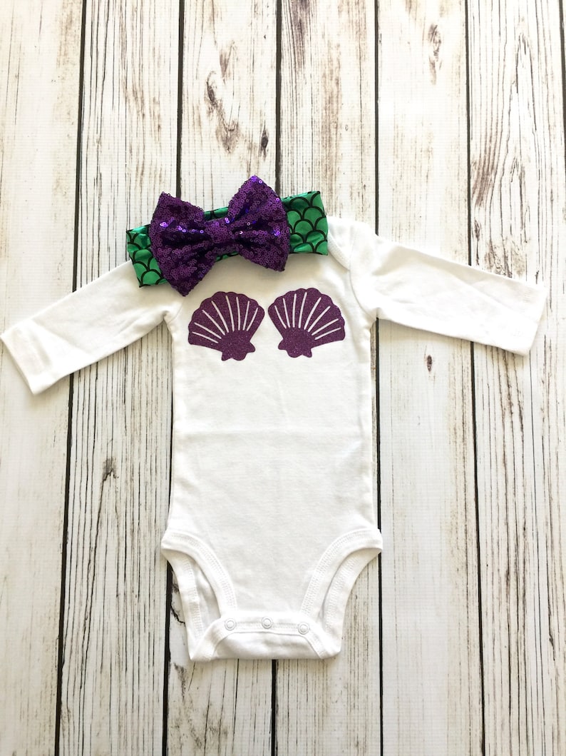 Peut inclure: Combinaison blanche pour b&eacute;b&eacute; avec des coquillages violets sur le devant. Un bandeau &agrave; sequins vert et violet avec un n&oelig;ud est plac&eacute; sur la combinaison.