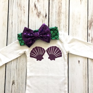 Peut inclure: Une grenouill&egrave;re blanche &agrave; manches longues avec un motif de coquillage en paillettes violettes. Un bandeau &agrave; sequins vert et violet avec un n&oelig;ud est inclus.