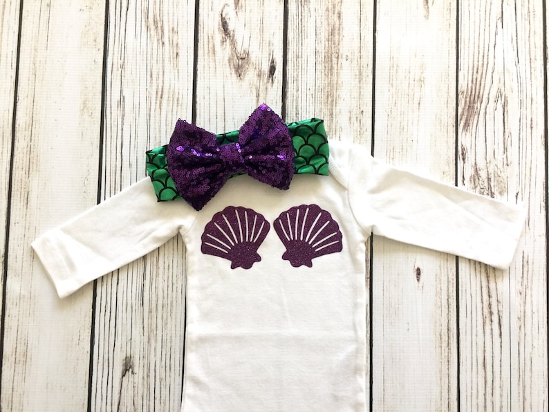 Peut inclure: Une grenouill&egrave;re blanche &agrave; manches longues avec un motif de coquillage en paillettes violettes. Un bandeau &agrave; sequins vert et violet avec un n&oelig;ud est inclus.
