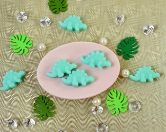 May include: Five green dinosaur-shaped soaps on a pink plate with green leaves and white pearls scattered around the plate on a gold background.