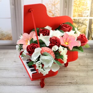May include: A red piano-shaped flower arrangement featuring a variety of blooms, including roses, lilies, and carnations. The piano is decorated with white and black keys.
