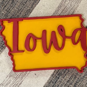 May include: A wooden cutout of the state of Iowa, with a yellow interior and a red outline. The word "Iowa" is written in red cursive script across the center of the state. The item is placed on a striped fabric background.