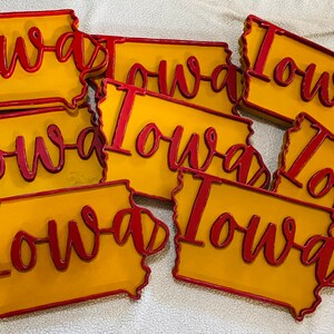 May include: A collection of Iowa state-shaped signs. Each sign features a yellow background with a red outline and the word "Iowa" in red cursive script. The signs are likely made of wood or a similar material.