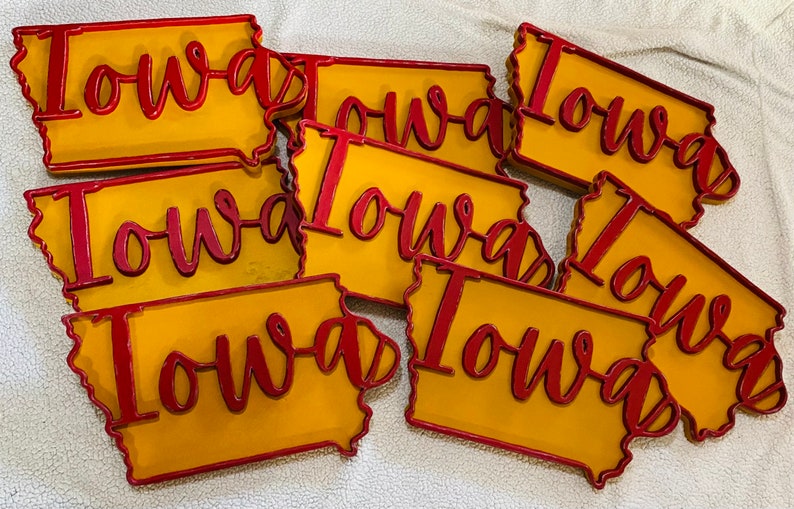 May include: A collection of Iowa state-shaped signs. Each sign features a yellow background with a red outline and the word "Iowa" in red cursive script. The signs are likely made of wood or a similar material.