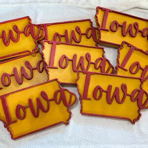 May include: A collection of wooden Iowa state-shaped signs. Each sign features a yellow background with a red outline and the word "Iowa" in red cursive lettering. The signs are arranged on a white surface.