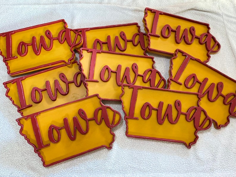 May include: A collection of wooden Iowa state-shaped signs. Each sign features a yellow background with a red outline and the word "Iowa" in red cursive lettering. The signs are arranged on a white surface.