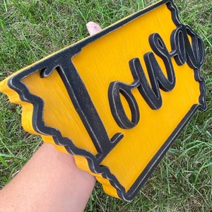 May include: A wooden sign in the shape of the state of Iowa, painted yellow with a black border. The word "Iowa" is written in a cursive font across the sign.