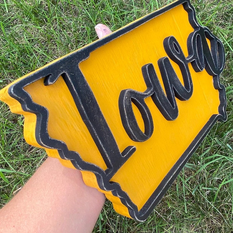 May include: A wooden sign in the shape of the state of Iowa, painted yellow with a black border. The word "Iowa" is written in a cursive font across the sign.