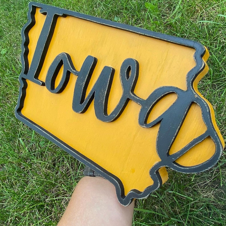 May include: A wooden sign in the shape of Iowa, painted yellow with a black outline and the word "Iowa" in a cursive font. The sign is held against a background of green grass.