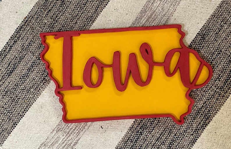 May include: A wooden cutout of the state of Iowa, with a yellow interior and a red outline. The word "Iowa" is written in red cursive script across the center of the state. The item is placed on a striped fabric background.