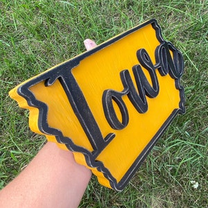 May include: Wooden sign in the shape of Iowa, painted yellow with a black border. The word "Iowa" is cut out in black script and mounted on the yellow background. The sign is held against a background of green grass.