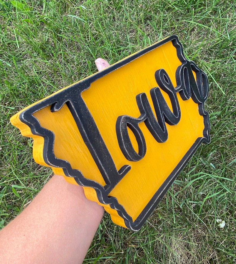 May include: Wooden sign in the shape of Iowa, painted yellow with a black border. The word "Iowa" is cut out in black script and mounted on the yellow background. The sign is held against a background of green grass.