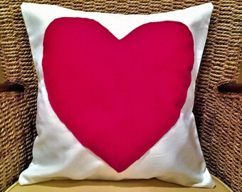 May include: A square decorative pillow with a large red heart shape on a white background. The pillow is placed on a wicker chair, creating a cozy and inviting aesthetic.