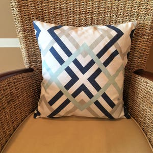 May include: Square decorative pillow with a geometric pattern in navy blue, light blue, and gray on a white background. The pillow is placed on a woven chair with a tan cushion.