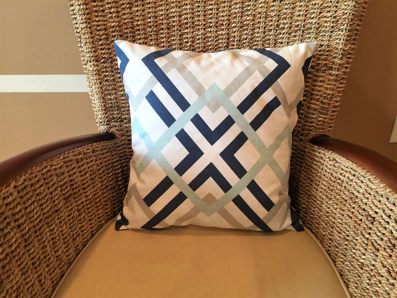 May include: Square decorative pillow with a geometric pattern in navy blue, light blue, and gray on a white background. The pillow is placed on a woven chair with a tan cushion.