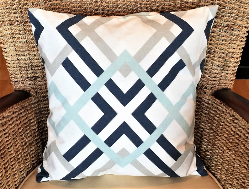 May include: Square throw pillow with a geometric pattern in navy blue, light blue, and gray on a white background. The pillow is placed on a wicker chair, showcasing its decorative design and color scheme.