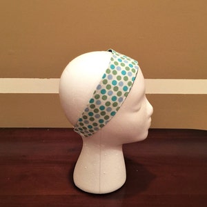 May include: A fabric headband featuring a pattern of blue and green polka dots on a white background. The headband is displayed on a white mannequin head. The headband is approximately 2.5 cm wide.